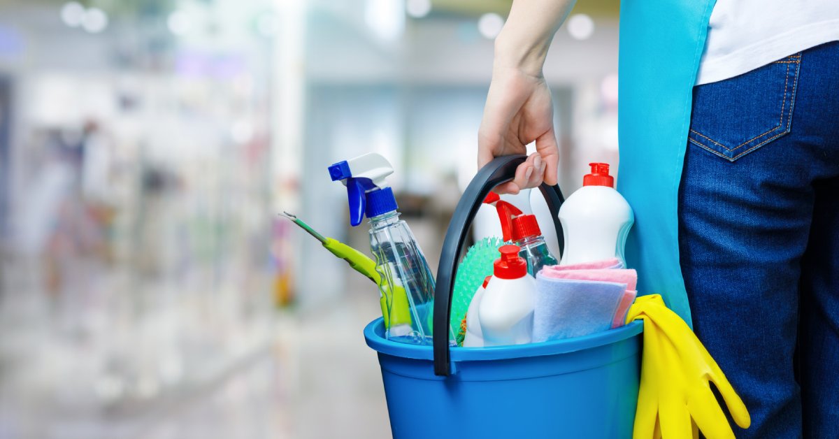 A woman's hand holding a plastic bucket full of cleaning supplies, such as brushes, cloths, and several bottles of cleaner.