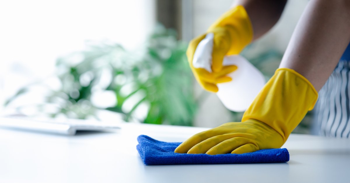 Two hands covered in yellow gloves cleaning a table's surface. One hand holds a cloth, the other holds a spray bottle.