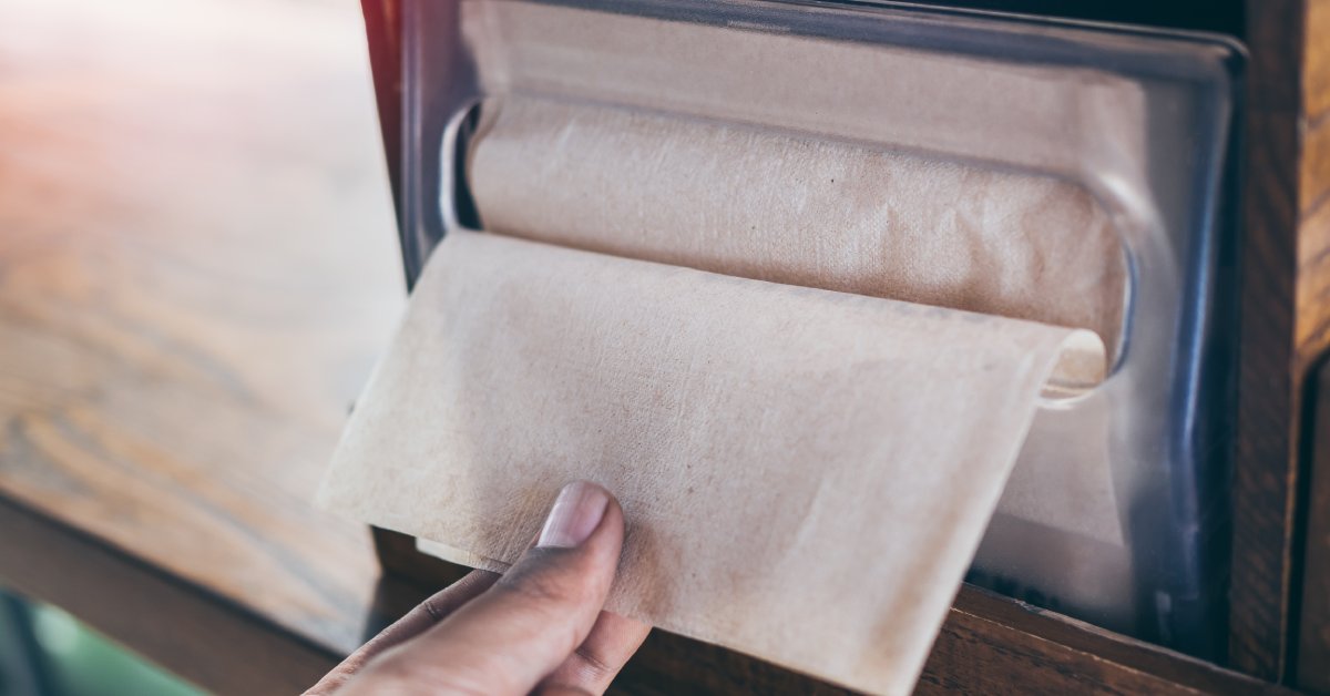 A person's hand grabbing a single brown, paper napkin from a napkin dispenser placed on a wooden countertop.