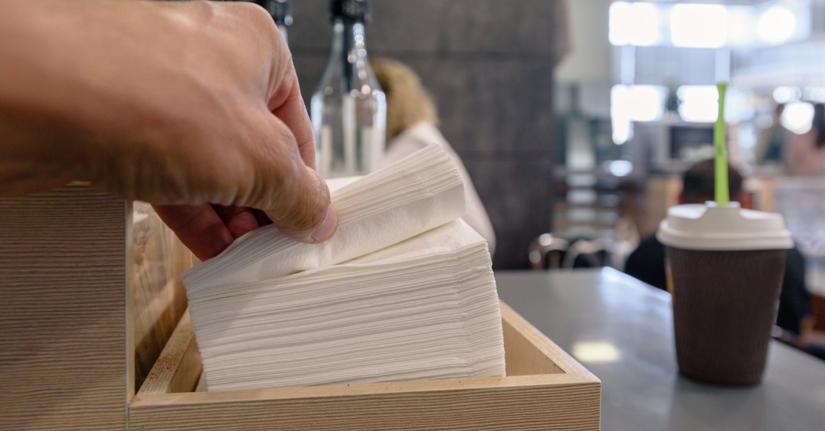 A closeup of a person's hands grabbing several napkins from a pile of many more at a condiment stand in a café.