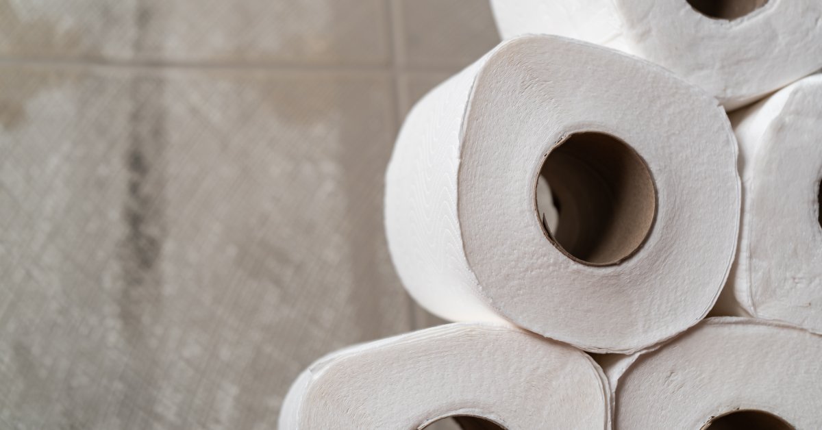 A close-up of a neatly arranged stack of standard toilet paper rolls. There is a tiled wall in the background.