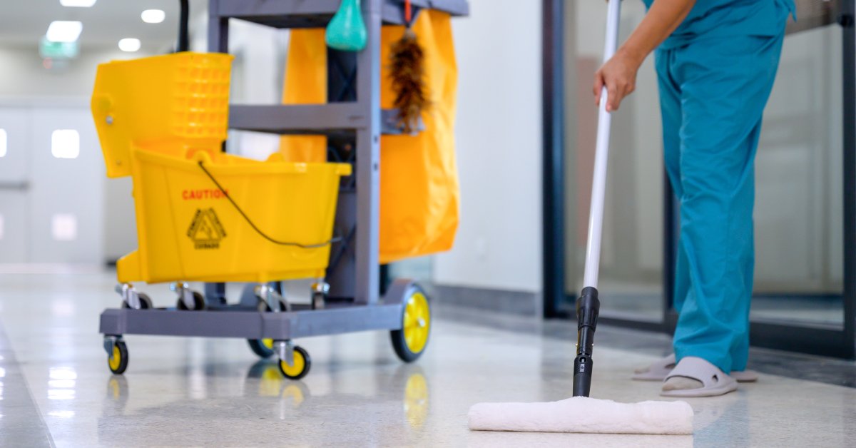 A person in a blue uniform mops a shiny epoxy floor with a yellow mop, using equipment from a nearby cleaning cart.