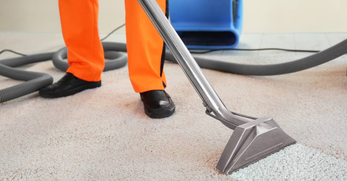 Worker wearing an orange uniform and black shoes using an extractor over a grey carpet, leaving a clean trail.