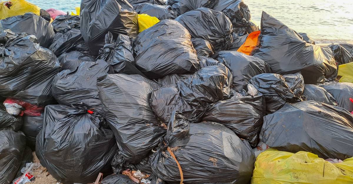 A large pile of plastic trash scattered along a sandy beach, all in different colored garbage bags with ties around the top.
