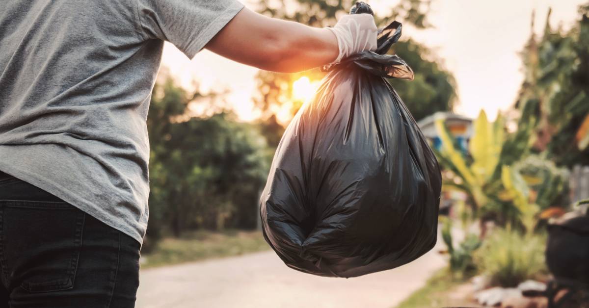 A woman holding a rolled-up garbage bag outside, and holding it over an outside garbage bin while the sun is setting.