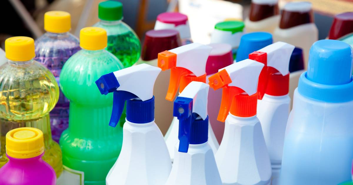 Multiple bottles of cleaning supplies, up close. The bottles are of different sizes, colors, and spray tops.
