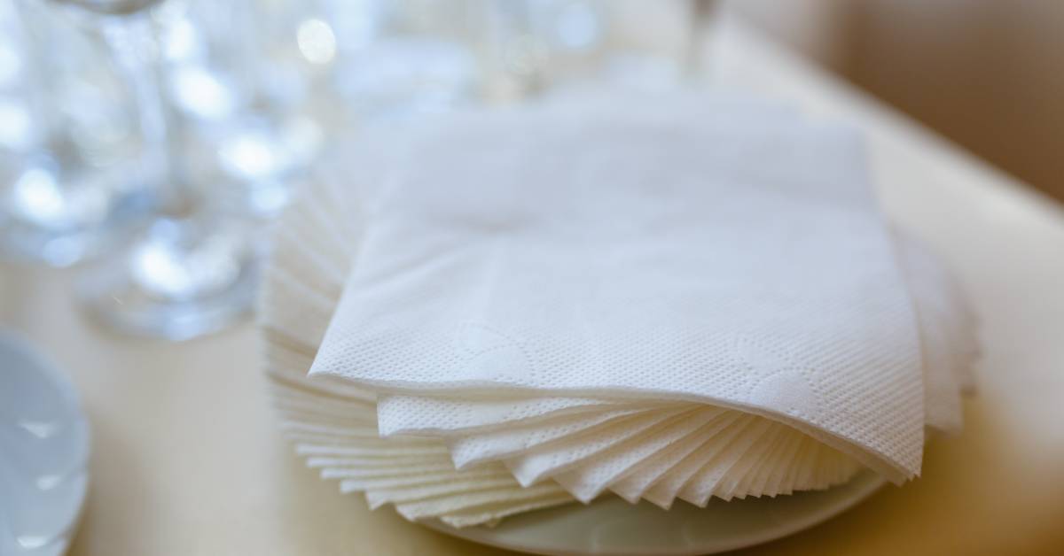 A stack of white paper napkins is placed on a small white dish over a table next to multiple blurred crystal glasses.