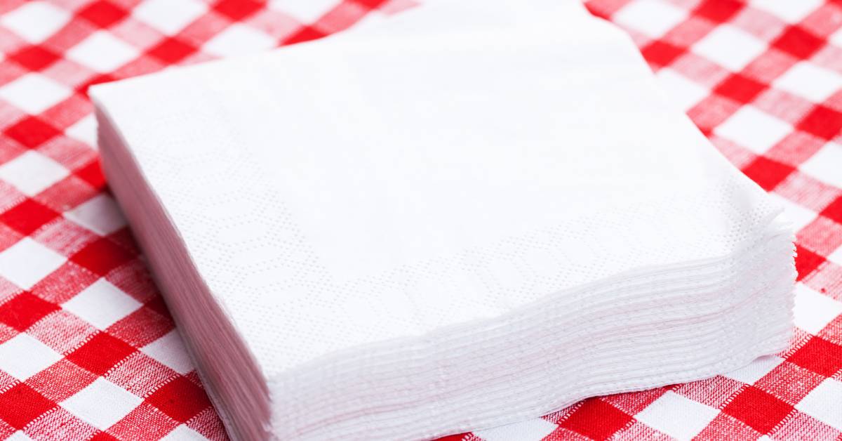 A stack of large white square napkins placed on top of a table with a white and red checkered picnic tablecloth.