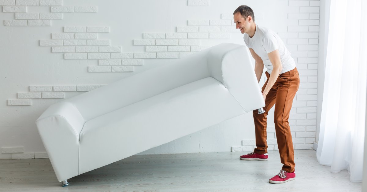 A man wearing brown pants and a white t-shirt lifting a long white couch inside a room with white floors.