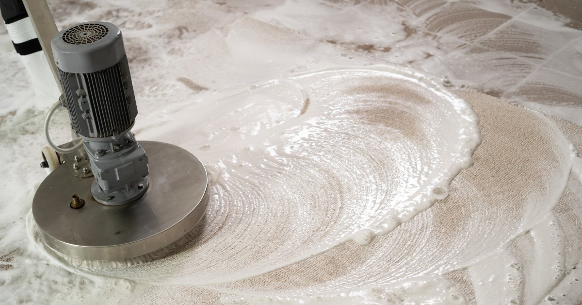 Close-up of the head of a large stainless steel scrubbing machine cleaning a tile floor and producing white foam.