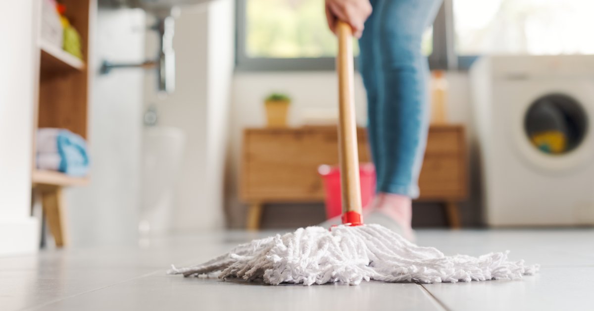 Close-up of the head of a mop with white strings held by a woman washing the white tiles of a bathroom floor.