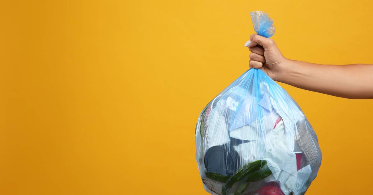 A person's arm holds a plastic blue trash bag filled with garbage in front of a dark yellow background.