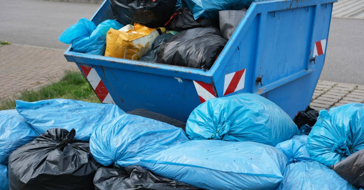 Multiple large blue and black plastic trash bags piled on top of each other on the ground in front of a container.