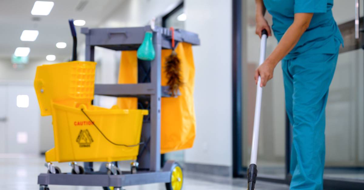 An employee wearing blue scrubs using a flat mop to clean the floor with a commercial cleaning cart parked behind them.