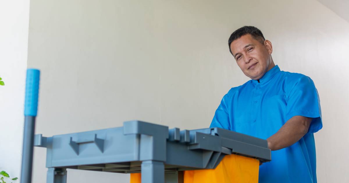 Man wearing blue scrubs pushing a cleaning cart with yellow fabric and a plastic grey structure down a hallway.