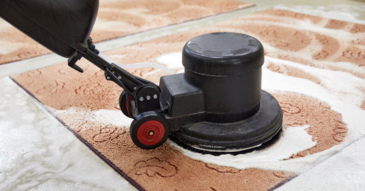 Black carpet cleaning machine with a rotary disk cleaning a brown rug with foam and water splashed across it.