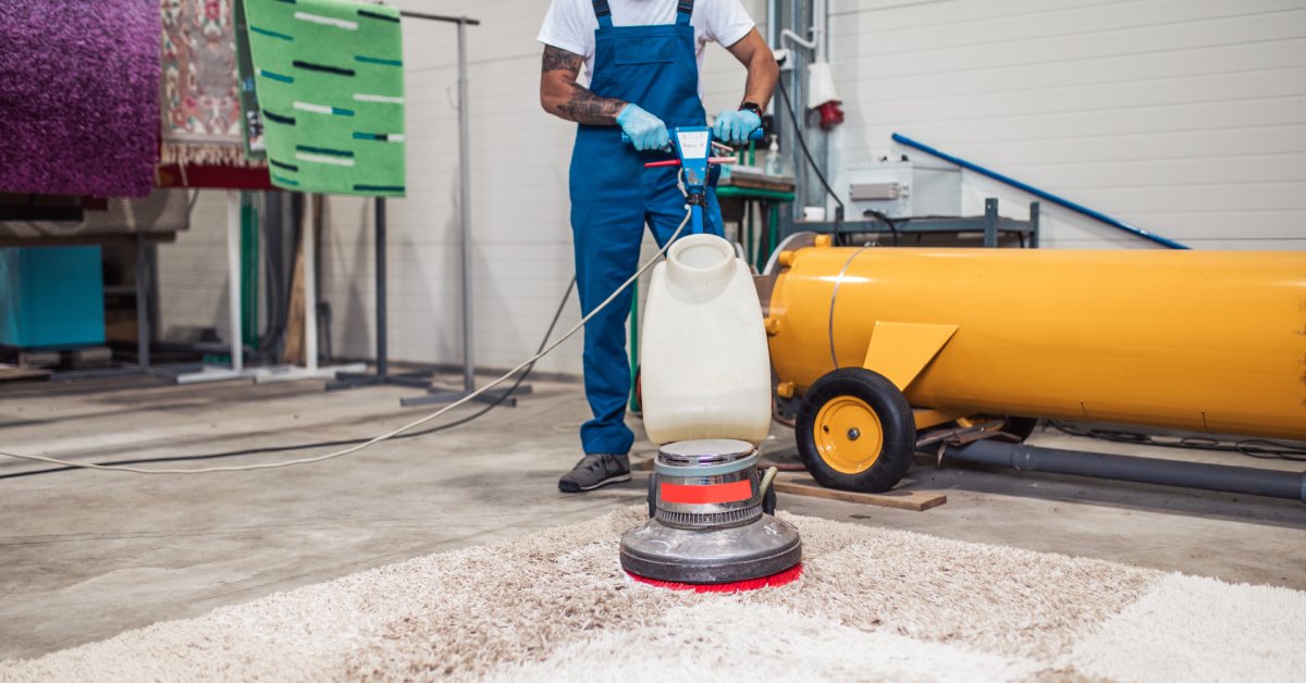 A worker wearing blue overalls using a cleaning machine over a large white rug inside an industrial environment.