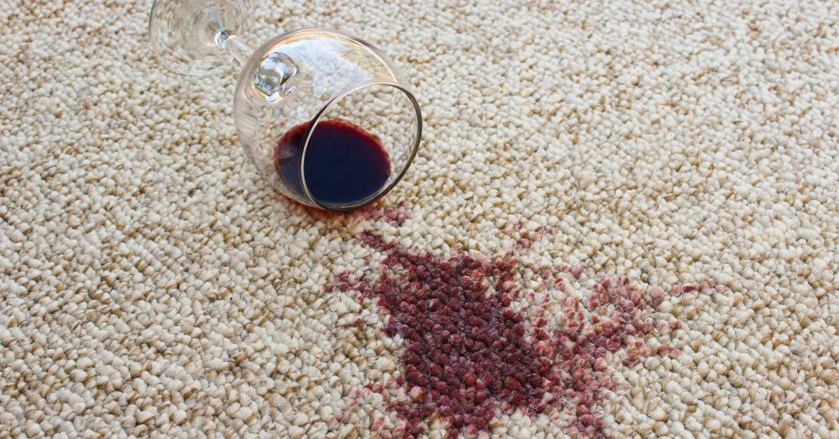 Knocked over glass with a small pool of red wine inside of it and a large red stain on a brown and white carpet.
