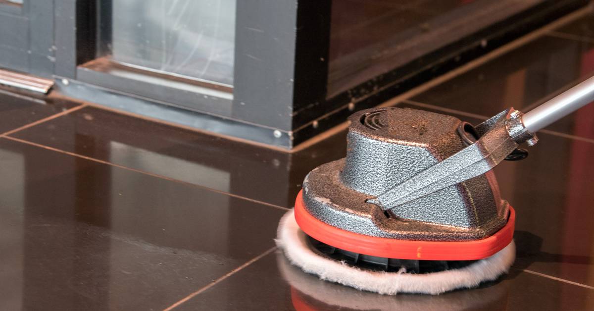 A close-up of the head of a gray and orange floor polisher cleaning dark tiled flooring near a corner.