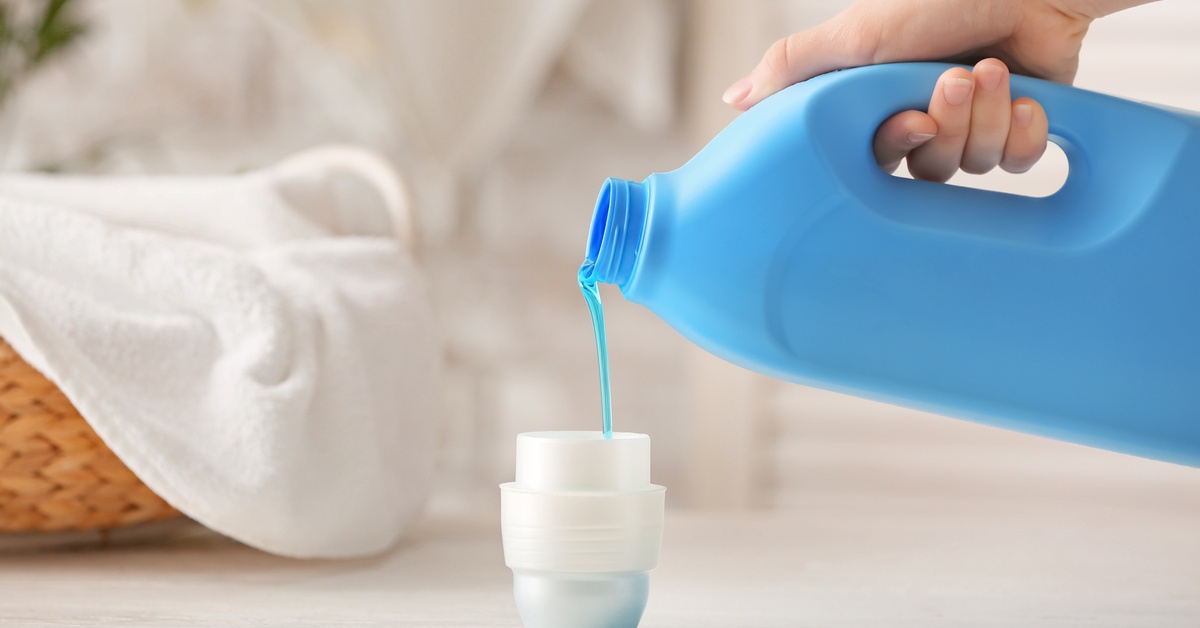 Close-up of a hand holding a large blue bottle pouring a thick liquid into a white cup over a white surface.