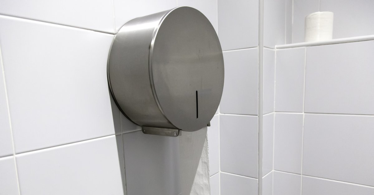 A large, circular toilet paper dispenser composed of metal is attached to a tile wall in a restroom.