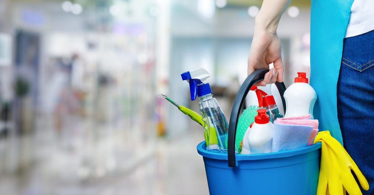 The back of a person standing and holding a blue bucket full of different-sized bottles of cleaning products.