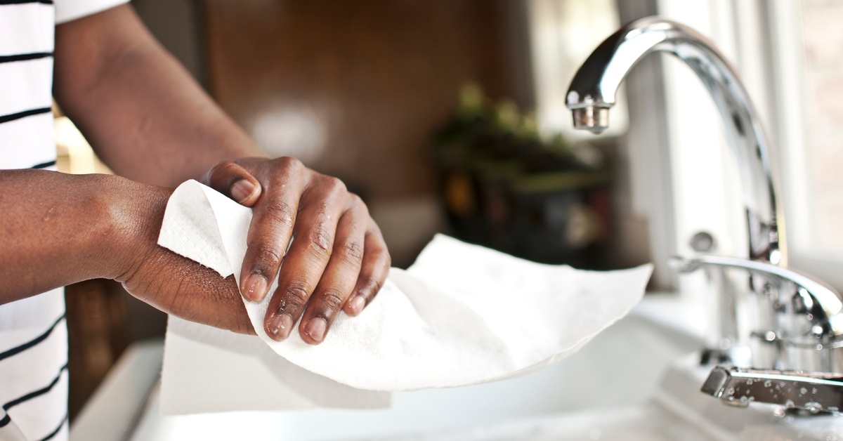 A person's hands, up close, using a white commercial-grade paper towel to dry them over a kitchen sink.